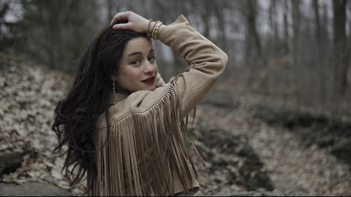 Amy has long, dark, curly hair with her hand in it. She is wearing a light brown top with a forest background and small, white flowers around her.