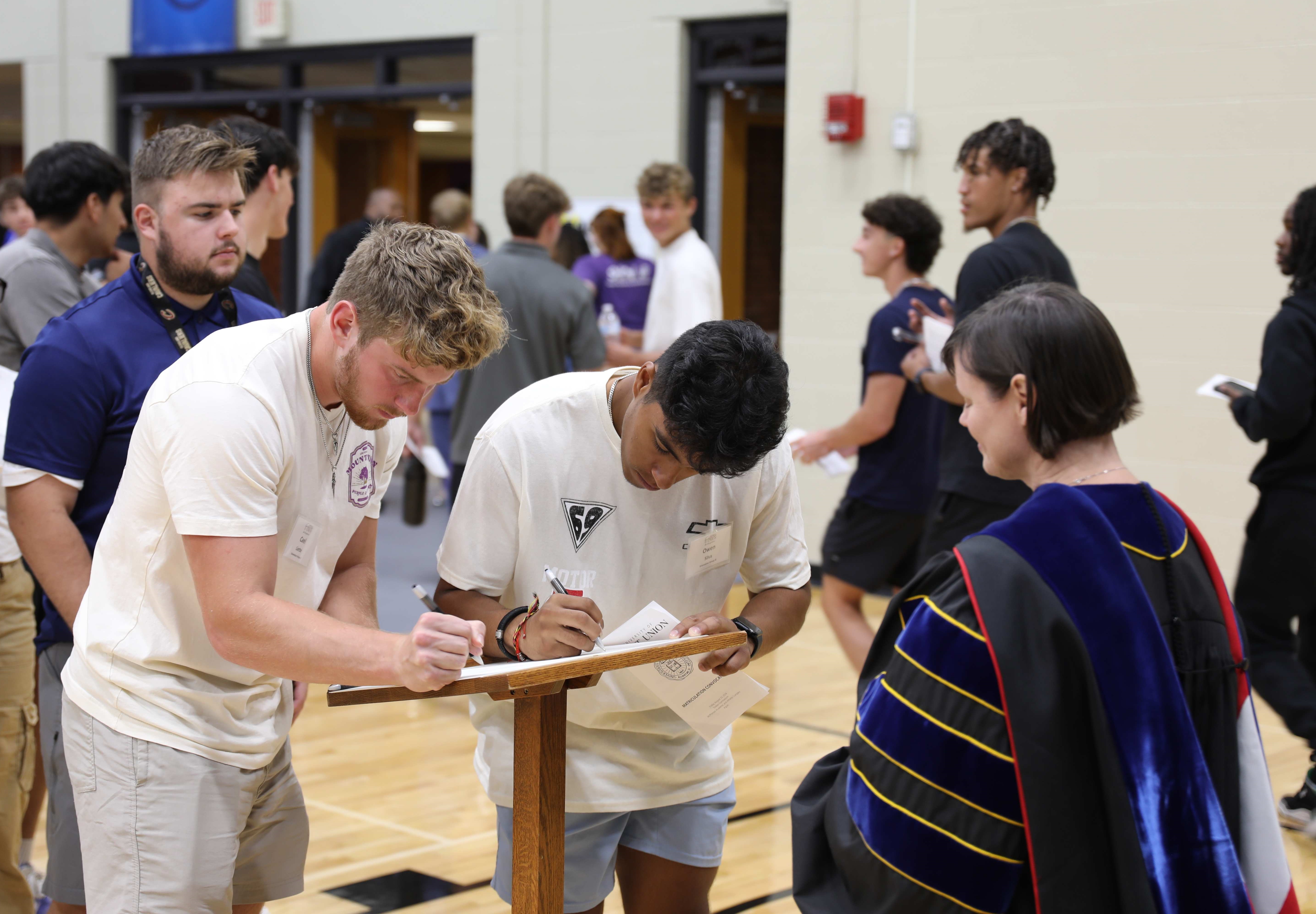 two students signing matriculation book as professor watches