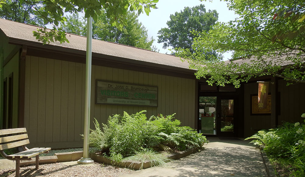 A photo of a building with a bench, flag pole, and flower beds at the Nature Center