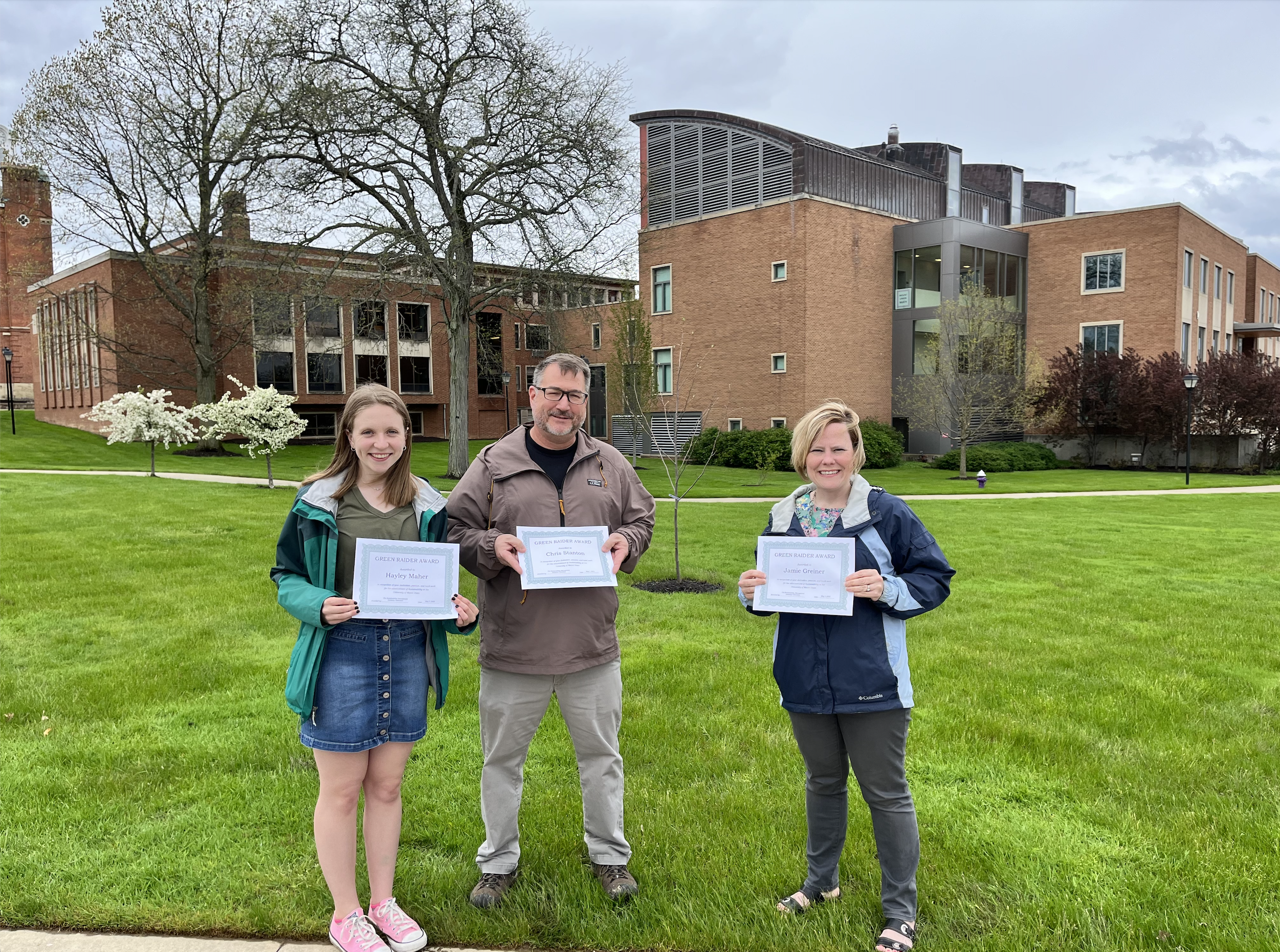 2022 Green Raider Awardees Hayley Maher '22, Dr. Chris Stanton, and Jamie Greiner stand next to a tree planted in their honor while holding their award certificates.